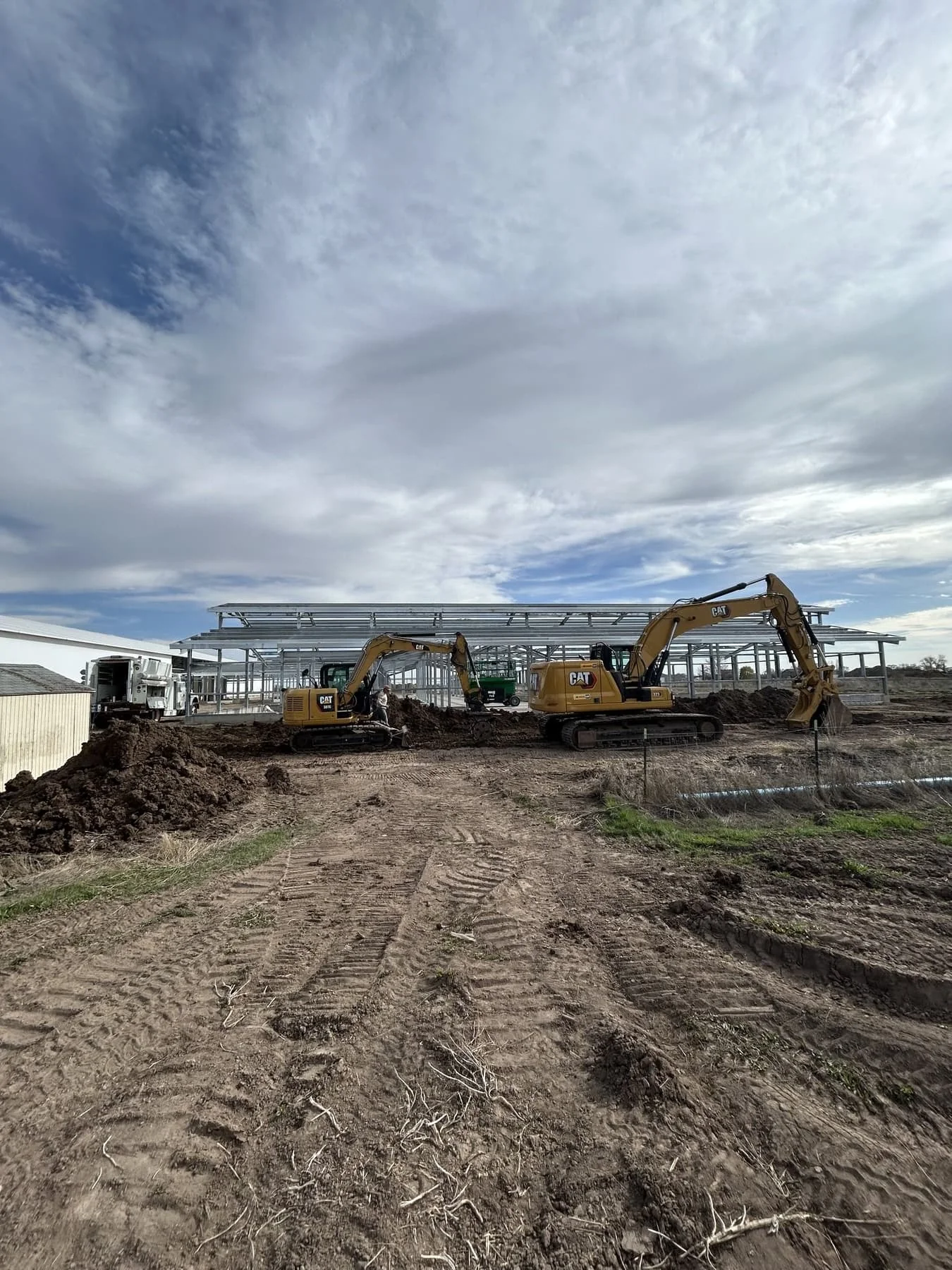 Two CAT excavators grading earth beside a large steel building frame under a dramatic cloudy sky